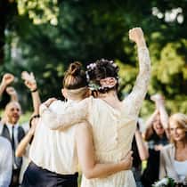 Was gilt für gleichgeschlechtliche Paare? Ein lesbisches Paar steht vor einer Hochzeitsgesellschaft im Freien.