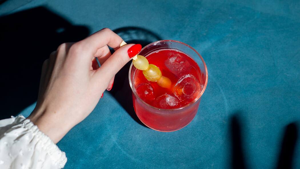 K.-o.-Tropfen im Glas: Wie schütze ich mich? eine manikürte Hand rührt in einem tiefen Cocktailglas, das eine knallrote Flüssigkeit enthält.