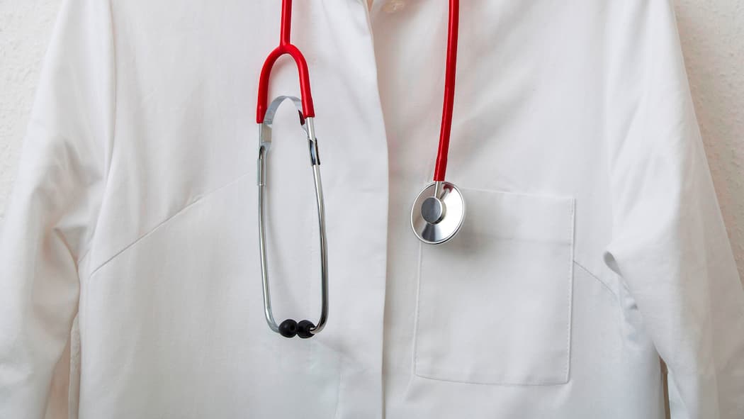 Hausarzt gewechselt – und plötzlich steigt die Prämie Doctor's coat hanging on a coat hanger on a coat rack, with a stethoscope