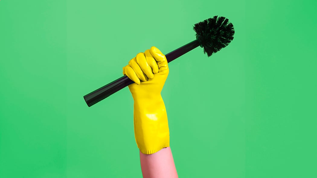 So melden Sie Hausangestellte an Ein Arm mit gelbem Handschuh hält eine schwarze Klobürste hoch vor grünem Hintergrund. Was gilt bei der Anstellung von Haushaltshilfen.