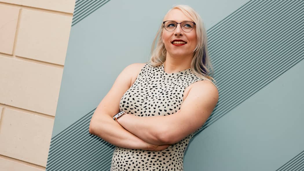 Endlich ich – eine trans Frau erzählt Die trans Frau Mia Macdonald trägt ein ärmelloses Kleid und steht selbstbewusst vor einer Wand. Sie lächelt und blickt nach rechts oben.