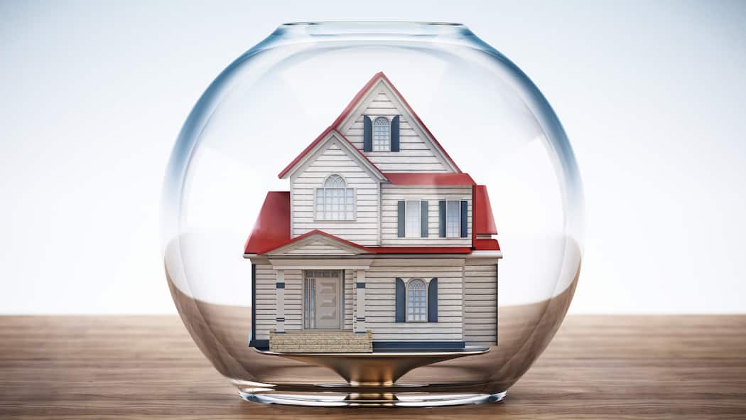 Wird meine Hypothek teurer, wenn die Immobilienblase platzt? Ein Haus steht auf einer hölzernen Oberfläche und wird von einem leeren Fischglas bedeckt, was einer Blase ähnlich sieht. Überbewertete Immobilienpreise könnten ebenfalls wie eine Blase platzen, sollten die Preise markant fallen.