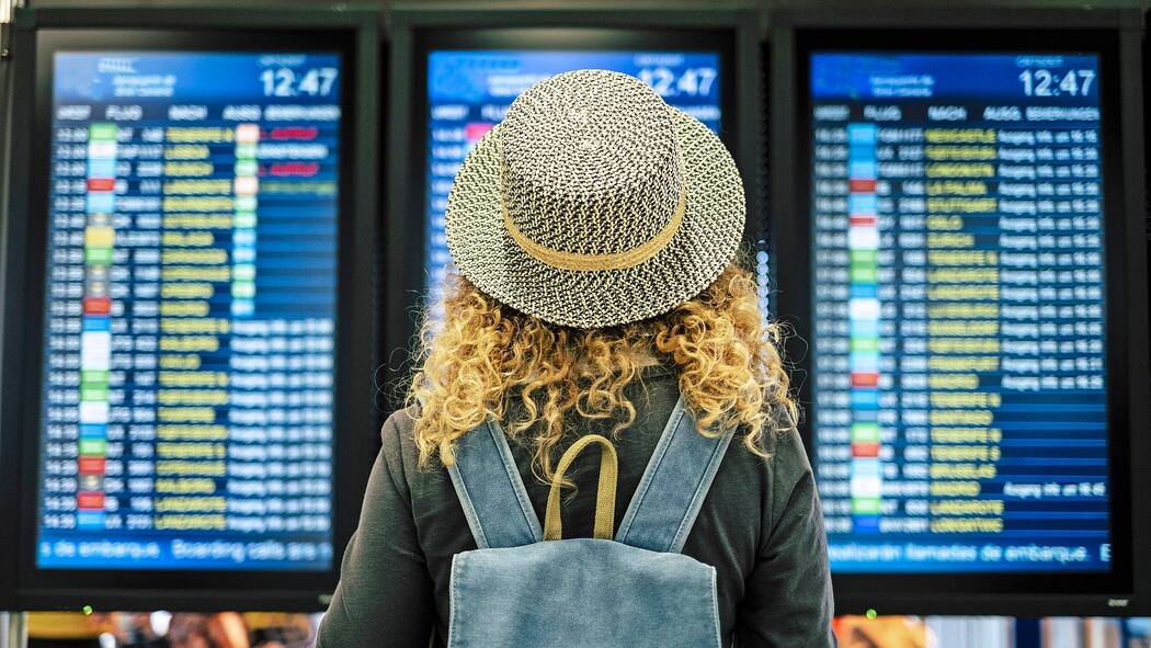 Wie bekomme ich mein Geld zurück? Eine Urlauberin mit Sommerhut steht vor mehreren Bildschirmen am Flughafen, welche die aktuellen Flüge anzeigen.