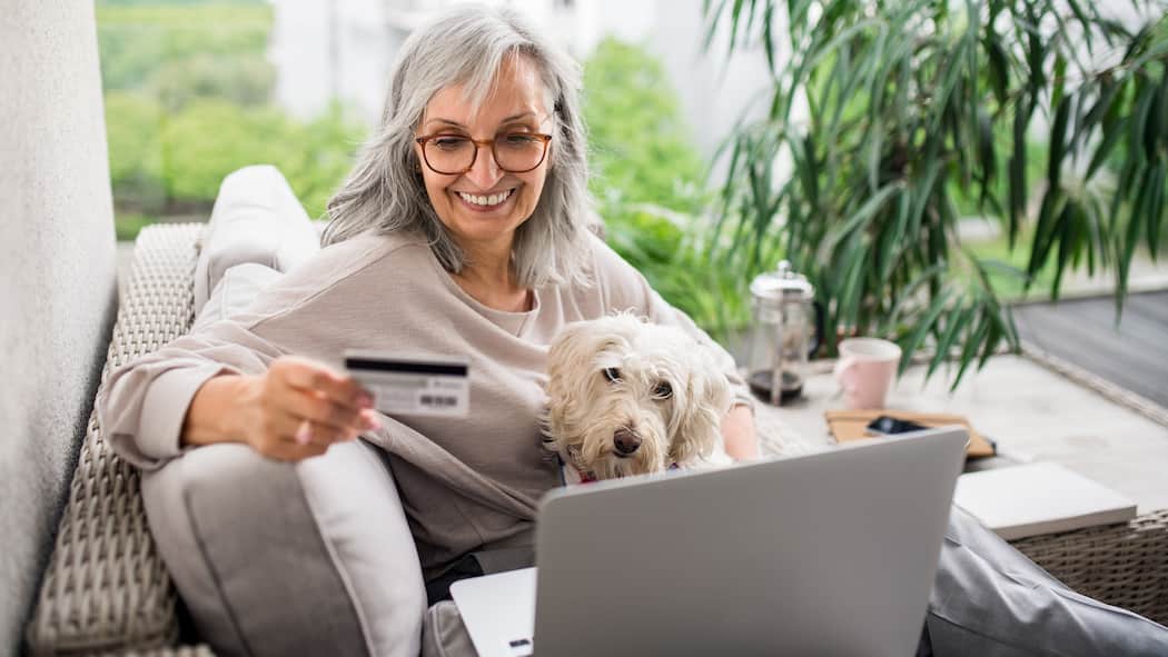 Soll ich auf E-Banking umsteigen? Eine ältere Frau sitzt in ihrem Wohnzimmer auf dem Sofa, ihr Hund auf dem Schoss. Ihr Blick ist auf ein Laptopbildschirm gerichtet, während sie in der anderen Hand eine Bankkarte hält.