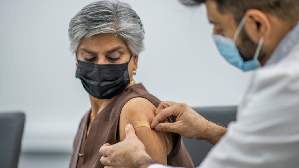 Wie gut und wie lange schützt die Corona-Impfung? Frau mit Schutzmaske bekommt Pflaster auf den Arm nach Corona-Impfung