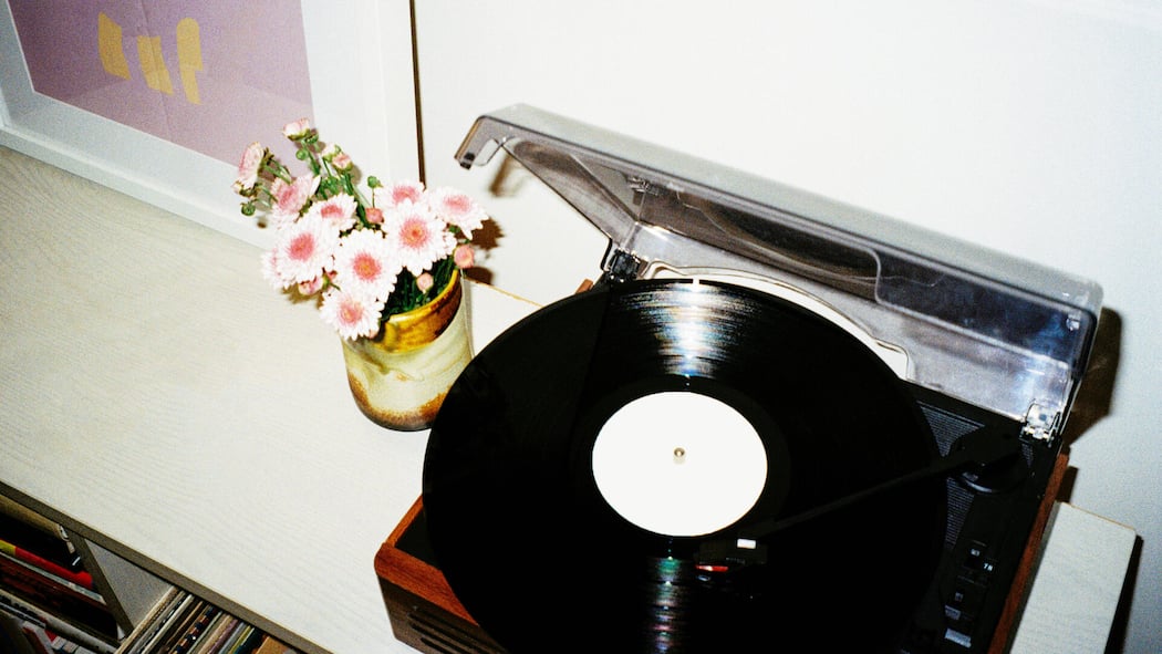 Als er das Lied vom Tod abspielte Ein Retro-Plattenspieler auf einem weißen Regal, aufgenommen vor einer weißen Wand. Neben dem Plattenspieler stehen eine kleine Vase mit rosa Blumen und ein gerahmtes Bild. Unter dem Regal sind Schallplatten und Bücher zu sehen.
