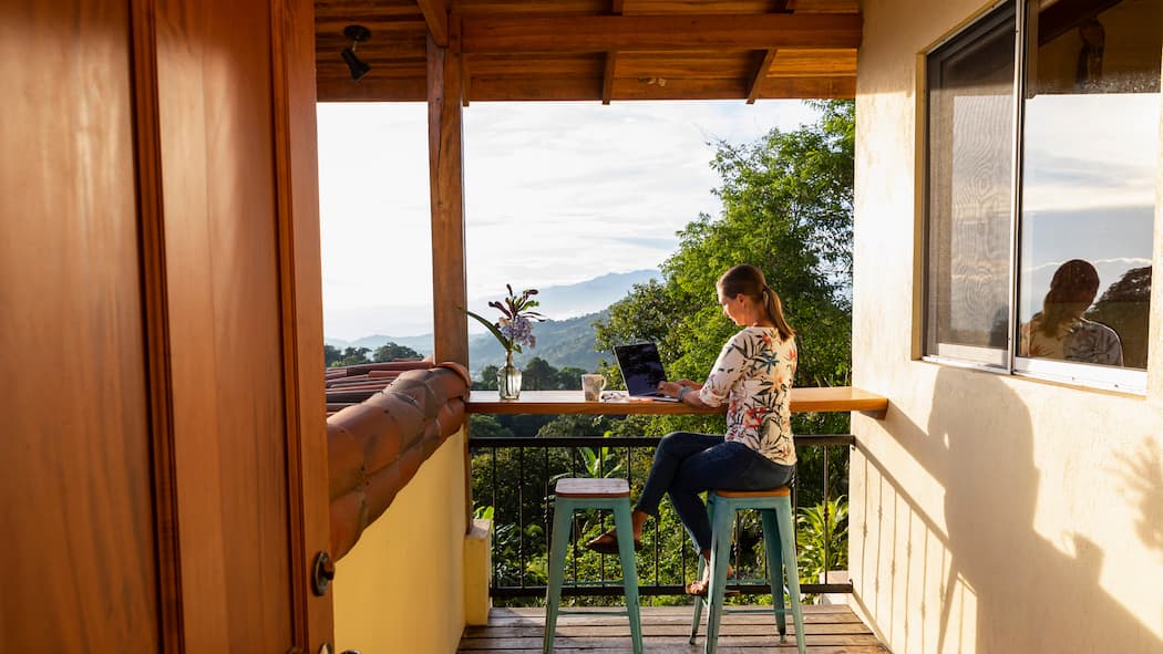 Wie man sein Büro an den Strand verlegt Eine junge Frau sitzt auf der Terrasse ihrer Wohnung, die sich inmitten einer grünen Landschaft befindet. Was können Remote Workerinnen tun, damit ihr Arbeitsaufenthalt im Ausland klappt und das auch noch nachhaltig für Land und Leute?