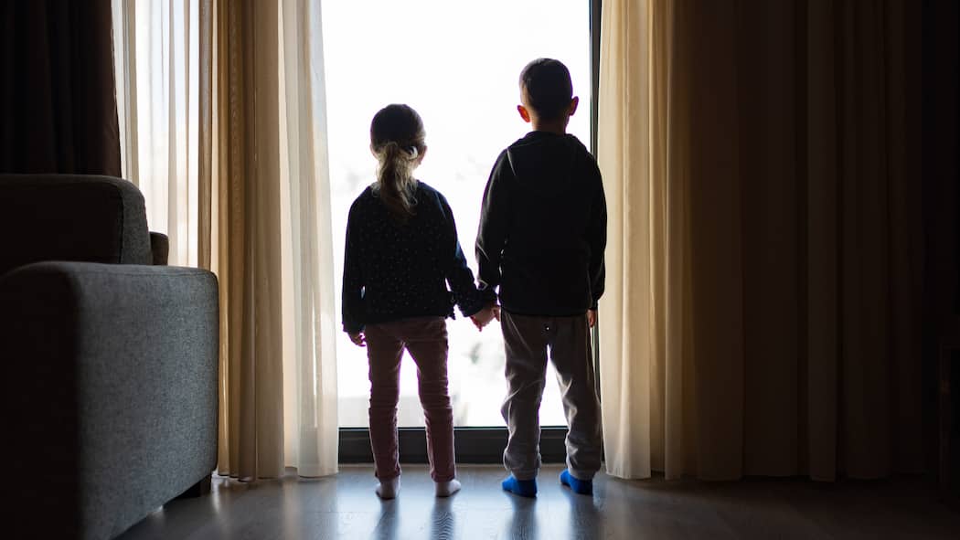 Wer wird sich um mein Kind kümmern? Zwei kleine Kinder stehen in einem verdunkelten Zimmer am Fenster, schauen durch das Fenster und halten sich die Hände.