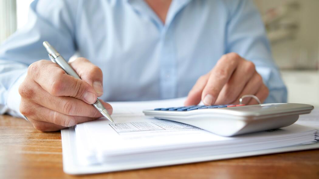 Den Geldfressern auf der Spur Eine Person sitzt am Schreibtisch und analysiert Zahlen. Daneben liegt ein Taschenrechner.