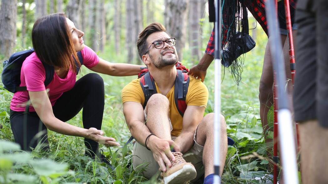 Tipps für eine sichere Wanderung Ein Wanderer mit Turnschuhen hat sich den Knöchel verstaucht, sitzt auf einem Waldboden und wird von vorbeilaufenden Wanderern angefragt, ob alles okay ist.