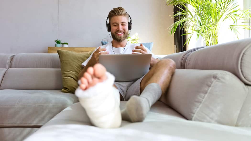 Mehr Rechte für Versicherte Ein junger Mann, dessen rechtes Bein mit einem Gips versehen ist, sitzt vergnügt auf dem Sofa während er einen Videoanruf mit seinem Laptop macht.