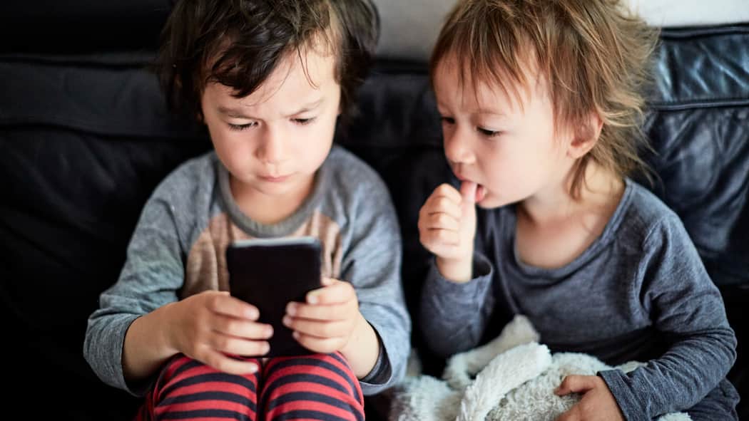 Kind kauft Spiele-Apps – müssen Eltern zahlen? Zwei kleine Jungs sitzen auf einem Sofa, der eine spielt mit dem Handy, der andere schaut ihm mit dem Finger nuckelnd dabei zu.
