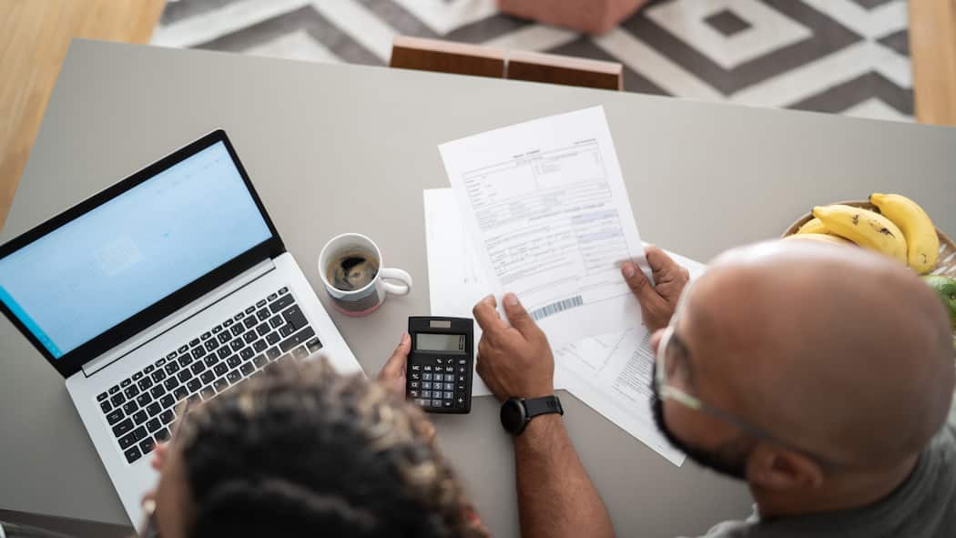 Bis wann eine Korrektur fordern? Ein Paar kontrolliert eine Rechnung mithilfe eines Taschenrechners und einem Laptop, der auf dem Küchentisch liegt.