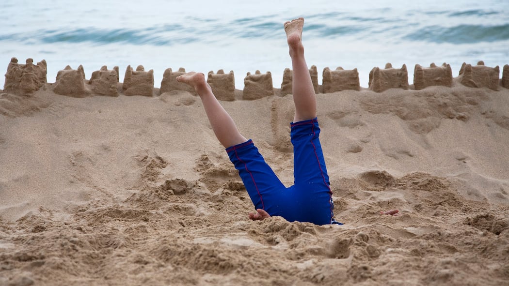 Müssen wir uns für die Reise ins Ausland extra versichern lassen? Eine Person steckt kopfvoran im Sand fest – Wer während der Ferien im Ausland erkrankt oder verunfallt, braucht in der Regel keine zusätzliche Reiseversicherung. Diese lohnt sich nur, falls man sich speziell absichern will oder bei Ländern mit hohen Behandlungskosten