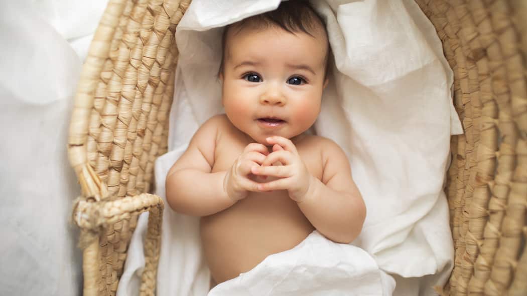 Auf Umwegen zum Baby Ein kleines Baby liegt umwickelt in weissen Laken in einem Korb und lächelt süss in die Kamera.