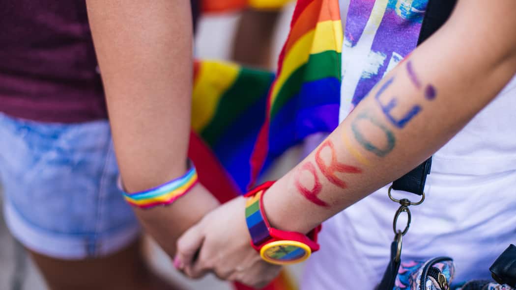 Was bedeutet LGBTQIA+? Ein Pärchen an einer Demonstration für die Rechte der LGBT-Community hält sich die Hände, im Hintergrund ist eine Regenbogenflagge zu erkennen. Wofür steht die Abkürzung «LGBTQIA+» und was gilt bezüglich dem Diskriminierungsverbot?