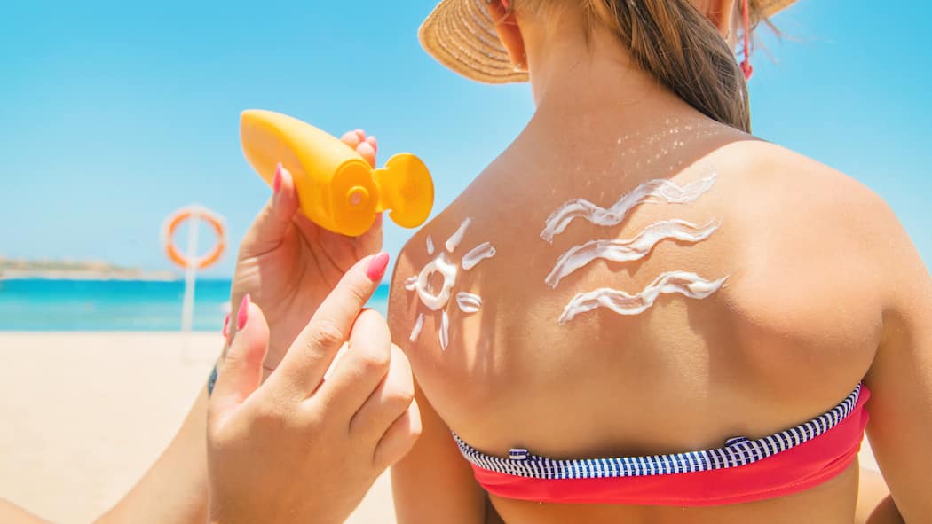 So vermeiden Sie hormonaktive Stoffe Ein kleines Mädchen am Strand steht mit dem Rücken zu ihrer Mutter, die ihr Sonnencreme in lustiger Wellen- und Sonnenform aufträgt.