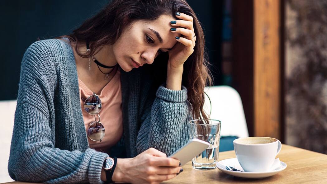 Von heute auf morgen ignoriert Eine junge Frau sitzt in einem Café und starrt betrübt auf ihren Handybildschirm.
