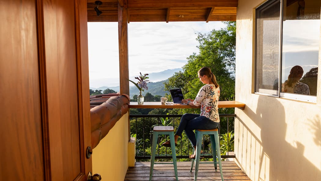 Das Büro sauber an den Strand zügeln Eine junge Frau sitzt auf der Terrasse ihrer Wohnung, die sich inmitten einer grünen Landschaft befindet. Was können Remote Workerinnen tun, damit ihr Arbeitsaufenthalt im Ausland klappt und das auch noch nachhaltig für Land und Leute?