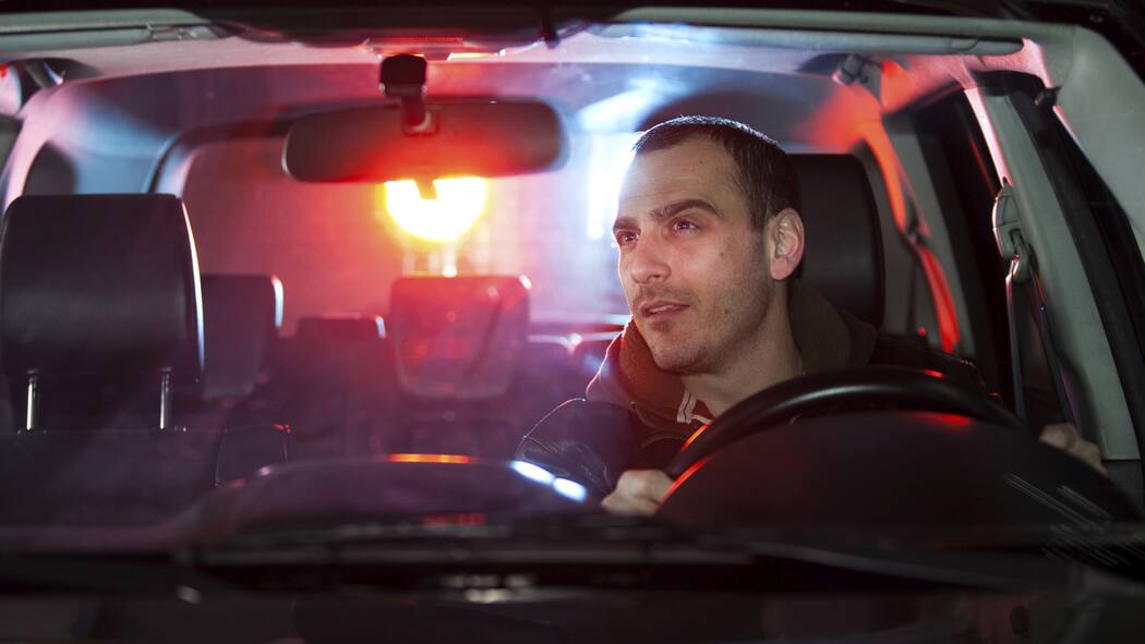 So schnell ist das Billett weg Ein junger Mann sitzt nachts am Steuer seines Auto, hinter ihm ist ein rot-blaues Licht eines Polizeifahrzeugs zu sehen.