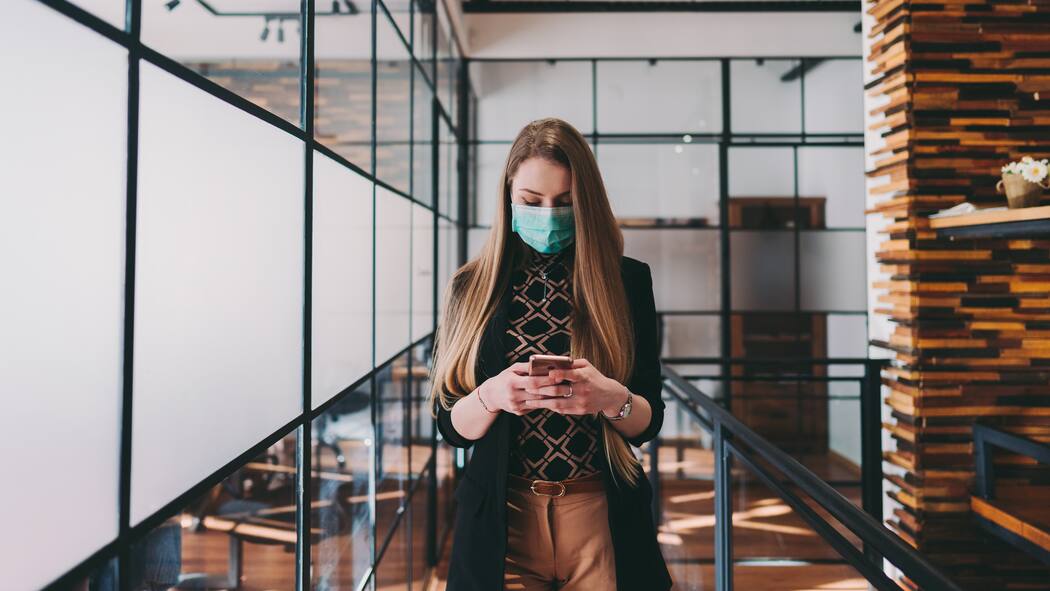 Darf ich trotz Verbot ins Büro? Eine Angestellte mit Schutzmaske geht durch ein Grossraumbüro und tippt gleichzeitig auf ihr Smartphone.