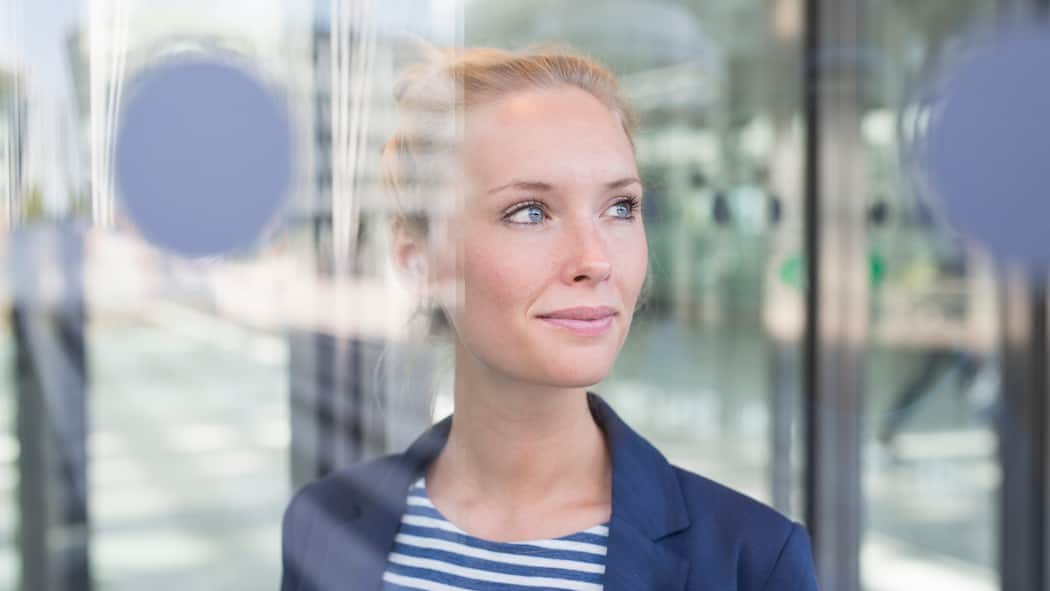 Selber entscheiden Eine Geschäftsfrau mit blauem Blazer schaut zufrieden und selbstbewusst durch ein verglastes Bürogebäude.