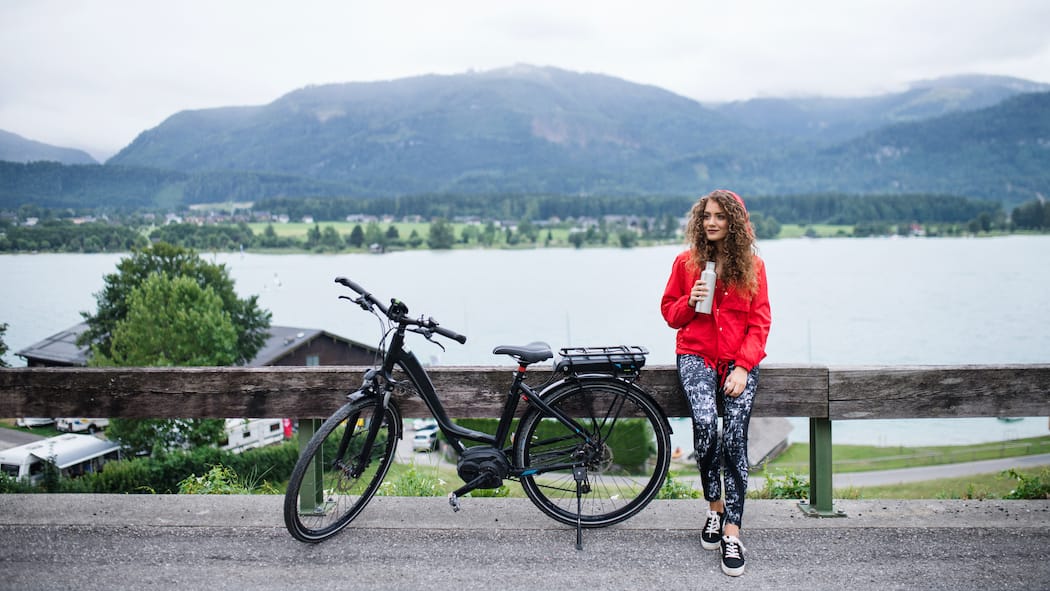 Worauf beim Kauf aus zweiter Hand achten Eine junge Frau sitzt bei ihrem Ausflug mit einem E-Bike auf einem Holzbacken am Strassenrand und trinkt während einer Pause von einer Wasserflasche.