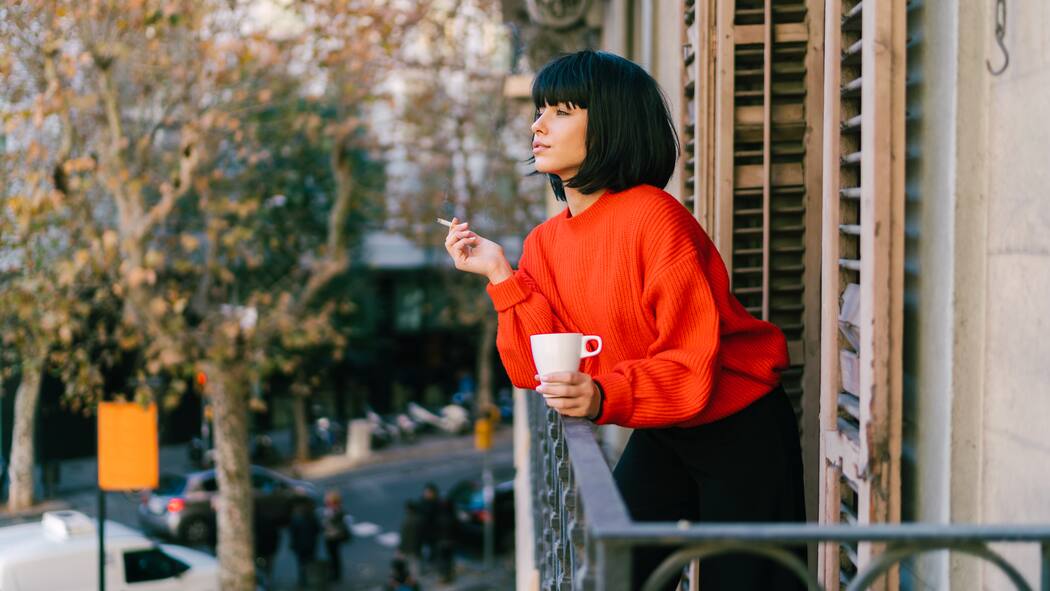 Wie mit schlechten Gewohnheiten aufhören? Eine junge Frau steht auf ihrem Balkon während einer Arbeitspause, die sie mit einer Tasse Kaffee und einer Zigarette verbringt.