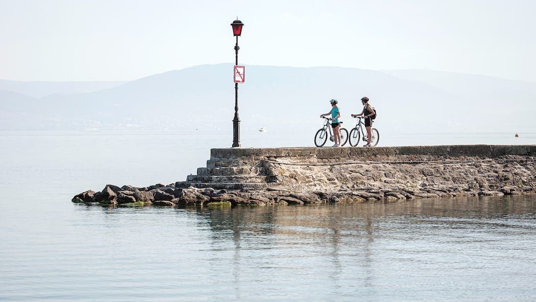 Im Genusstempo durch die Schweiz Zwei Velofahrerinnen befinden sich auf einem Steg in Estavayer-le-Lac und blicken hinaus auf den Neuenburgersee.