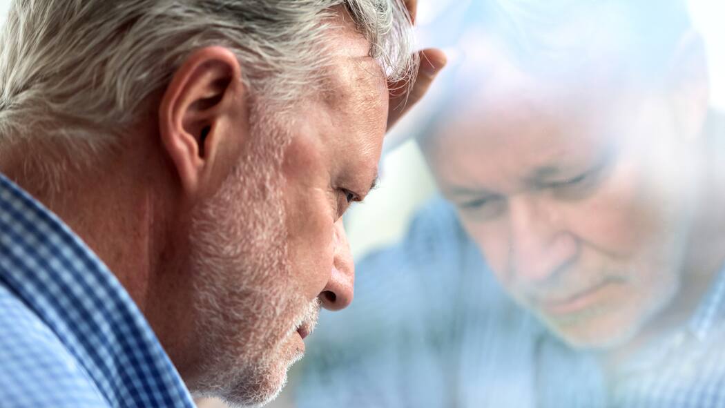 Schmerzhaftes Nein Grauhaariger Mann legt nachdenklich seinen Kopf an eine Scheibe und spiegelt sich darin.