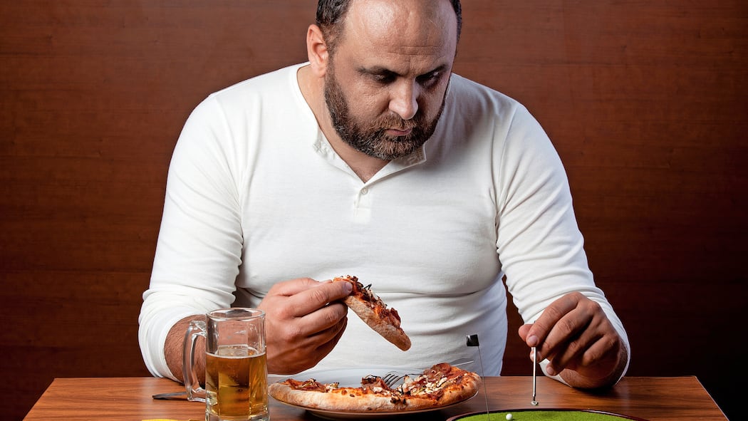 So entlasten Sie Ihre Leber Ein übergewichtiger Mann isst eine Pizza und spielt Tischgolf, um sich nicht bewegen zu müssen.