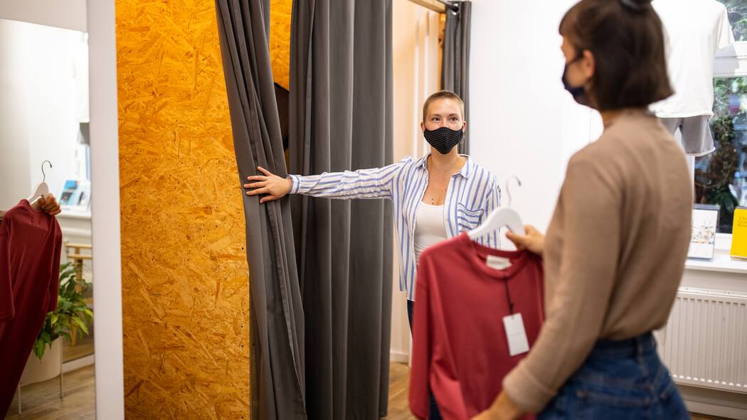 Kann mich der Chef einfach heimschicken? Eine Angestellte in einem Kleiderladen ist einer Kundin behilflich und öffnet für sie den Vorhang für die Umkleidekabine.