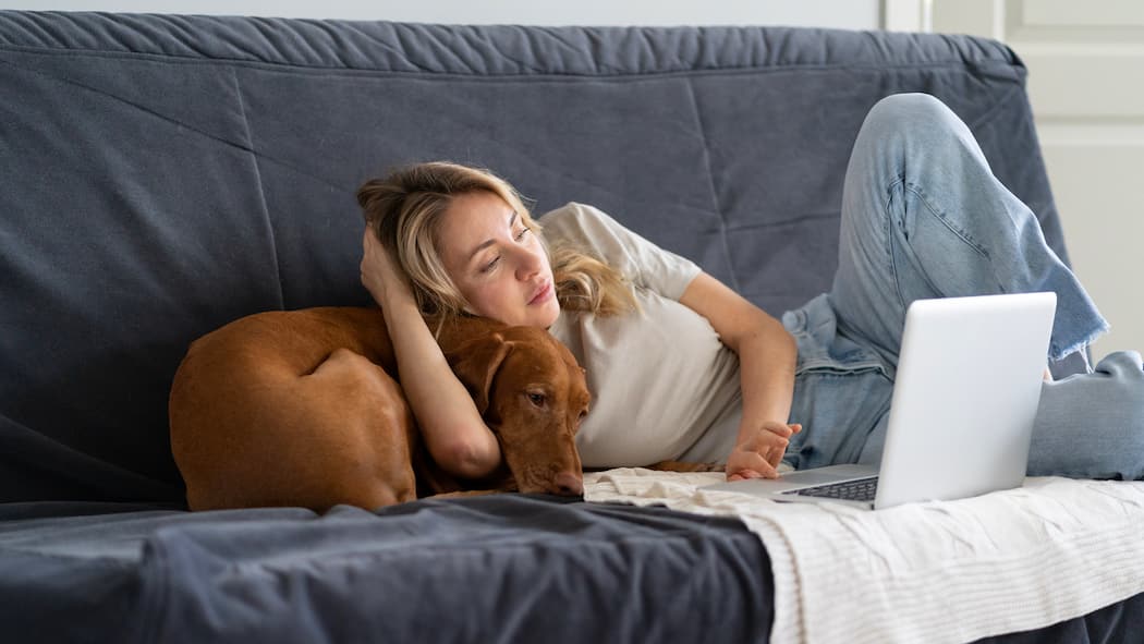 Wieso fühlen wir uns ständig müde? Eine junge Frau liegt auf dem Sofa mit ihrem Hund und fällt demnächst vor ihrem Laptop in den Schlaf.