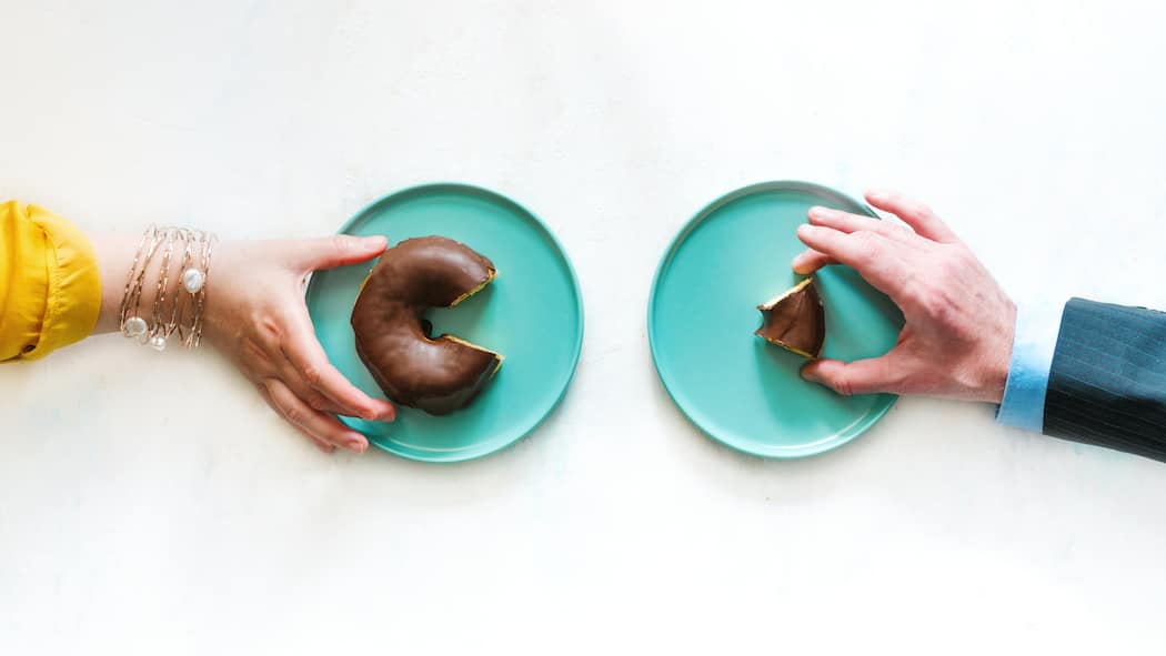 So holen sich Frauen mehr Zwei verschiedene Hände greifen nach zwei unterschiedlich grossen Donuts. Die Hand der Frau hat ein grösseres Donutstück als die Hand des Mannes.