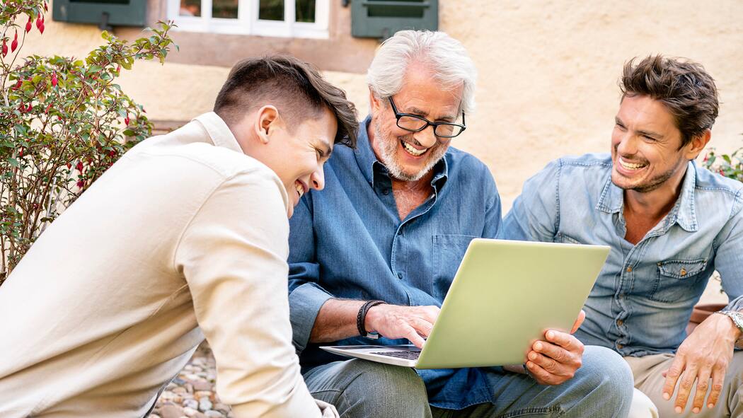 Soll ich Rechnungen mit E-Bill bezahlen? Ein Vater sitzt mit seinen beiden erwachsenen Söhnen im Garten seines Hauses und hat einen Laptop bei sich.