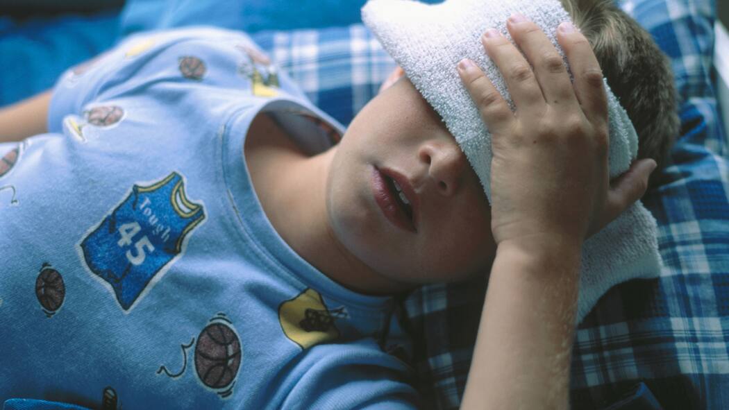 Jedes zehnte Kind hat Migräne Ein Junge liegt im Bett und hält sich einen Waschlappen vor die Augen.