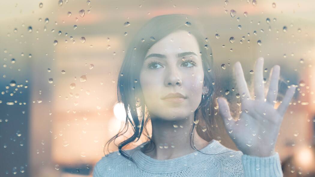 «Ich fühle mich so ausgelaugt» Eine junge Frau blickt in Gedanken versunken aus dem Fenster und hält ihre linke Hand gegen das Fensterglas, auf dem einzelne Regentropfen liegen.