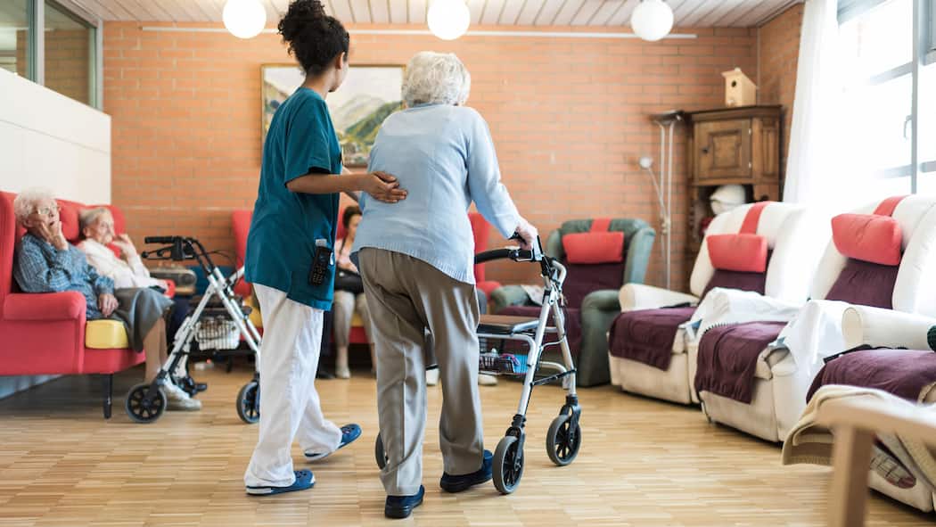 Worauf ist beim Umzug in ein anderes Pflegeheim zu achten? Pflegerin stützt alte Frau mit Rollator im Altersheim