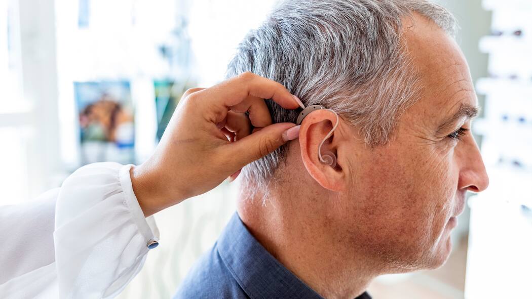 Ist mein Hörgerät bei Verlust versichert? Ein älterer Mann sitzt in einer Praxis. Die Ärztin hilft ihm dabei, sein Hörgerät anzupassen.