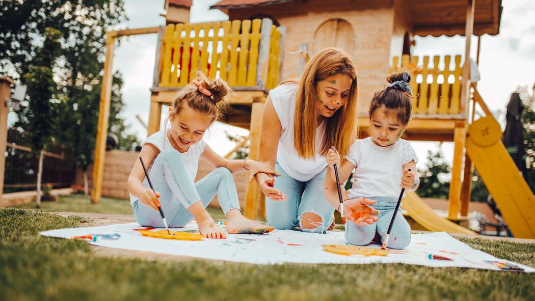 Das Abc des Spielens Zwei Kinder im Freien auf einer Wiese malen mit Farbe auf ein Blatt Papier und haben zusammen mit ihrer Mutter sichtlich Spass am Spielen.