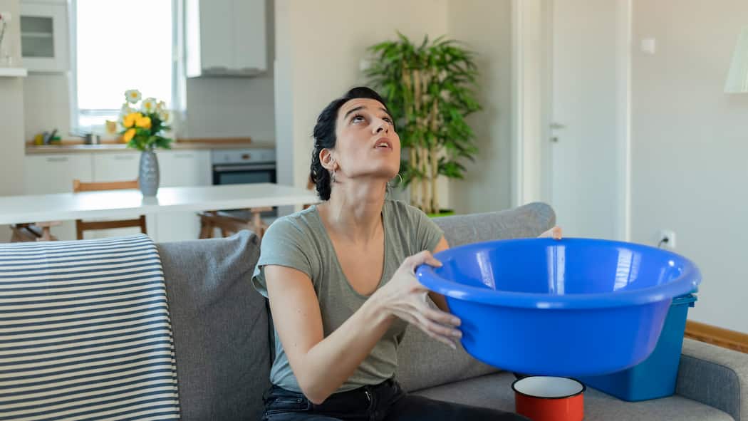 Jetzt aber hopp, Herr Vermieter! Eine Mieterin sitzt in der Wohnung auf ihrem Sofa und hält einen blauen Plastikeimer in den Händen, um Wasser von einem tropfenden Leck aufzufangen.