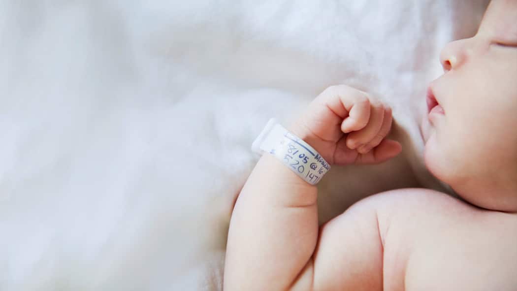 Wie ändert man den Familiennamen? Ein Neugeborenes liegt auf der Geburtsstation. Das Baby schläft tief und trägt am rechten Handgelenk einen Zettel mit Geburtsdaten.