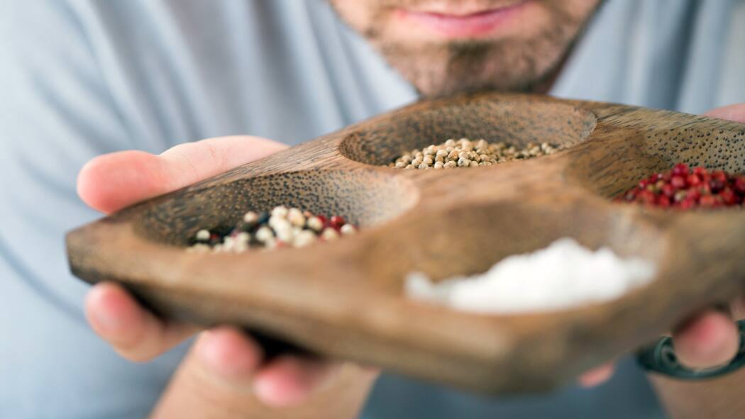 Heute schon am Pfeffer geschnuppert? Vier verschiedene Gewürze sind auf einem Holzteller verteilt. Ein Mann riecht an den Gewürzen.