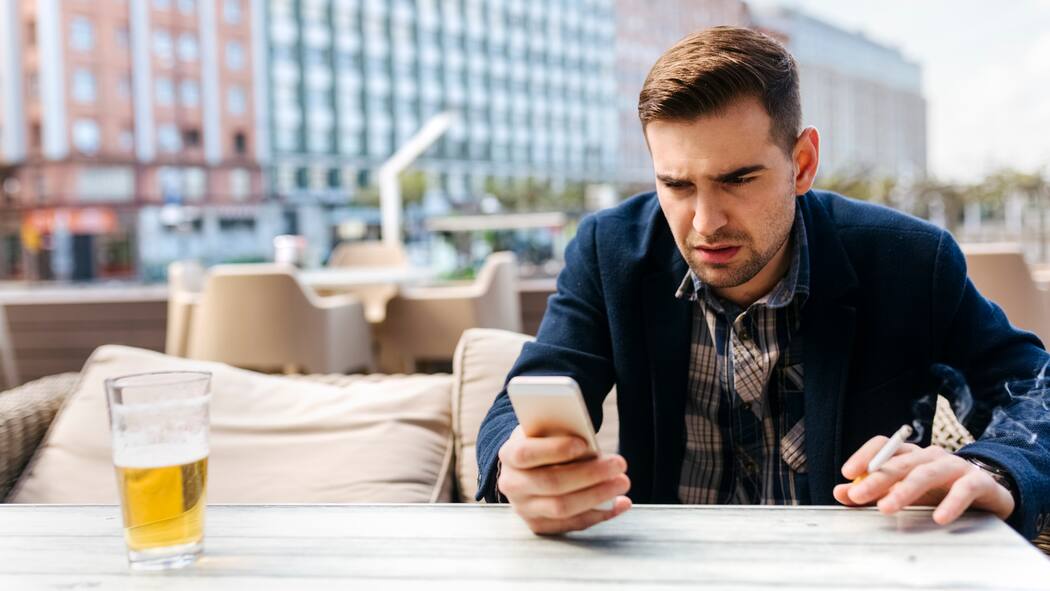Weniger Ärger mit dem Telefon Ein Mann sitzt auf einer Terrasse im Restaurant und schaut leicht genervt auf sein Handy während er eine Zigarette raucht.