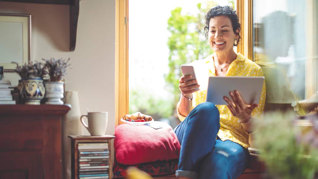Aus der Ferne auf mobile Geräte zugreifen Eine Frau sitzt entspannt zu Hause auf dem Fenstersims und hält in ihrer linken Hand ein Tablet, in der rechten ein Smartphone.