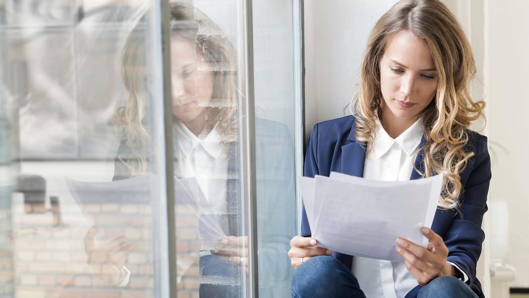 Weg vom Job, aber mit Zeugnis Eine Frau sitzt auf dem Fenstersims und liest ihr Zwischenzeugnis durch.