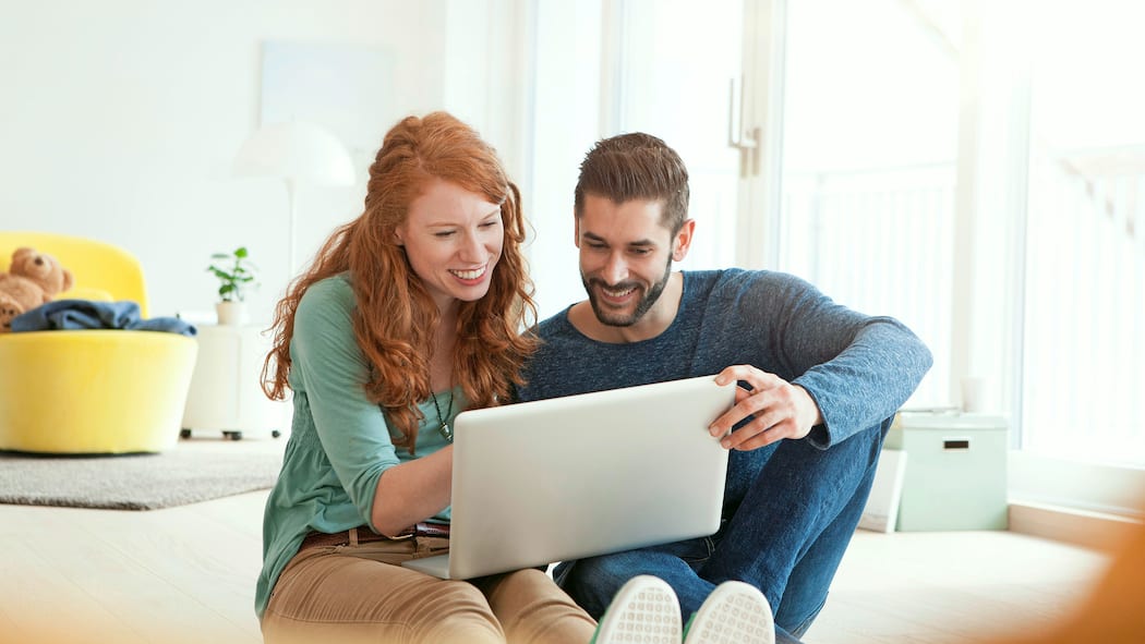 Vorsicht bei einer virtuellen Besichtigung Junges Paar sitzt zusammen auf dem Fussboden im Wohnzimmer und schaut sich auf dem Laptop etwas an.