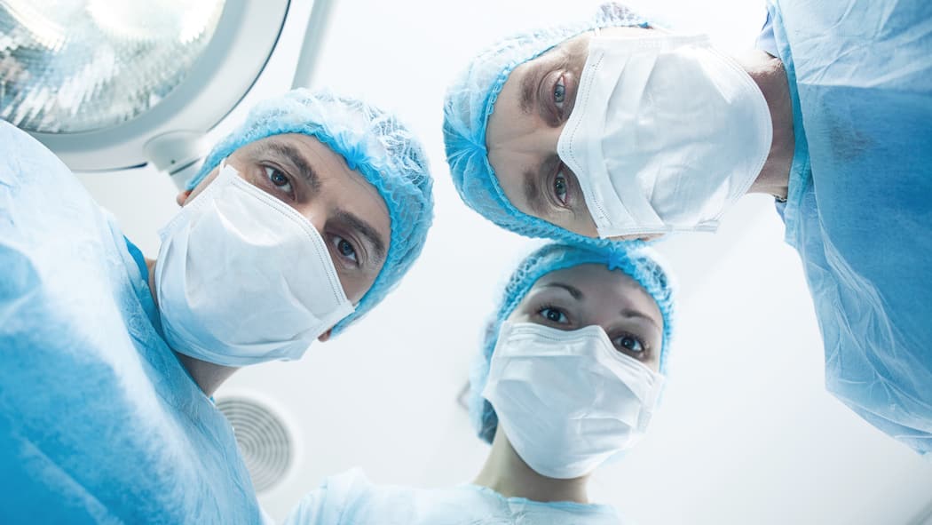 Verliere ich meinen neuen Job? Zwei Chirurgen und eine Chirurgin stehen in einem Operationssaal und blicken von oben auf den vermeintlichen Patienten herab.