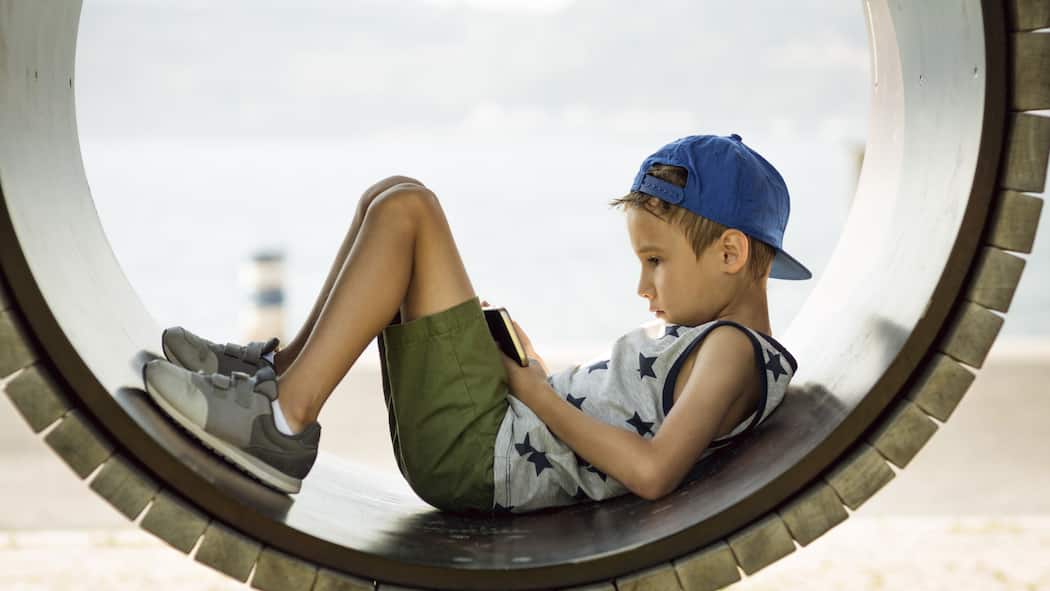 Wie weit sollen Eltern das Handy überwachen? Ein kleiner Junge auf einem Spielplatz hat seinen Blick auf ein Smartphone vertieft.