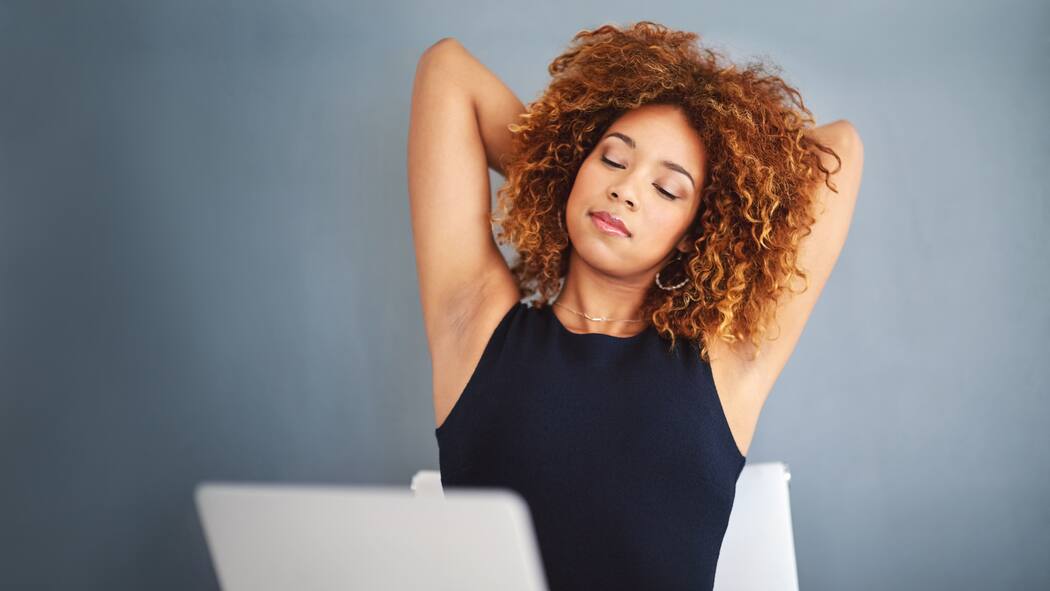 So kommt Bewegung ins Büro Eine Frau sitzt vor dem Laptop und streckt sich.